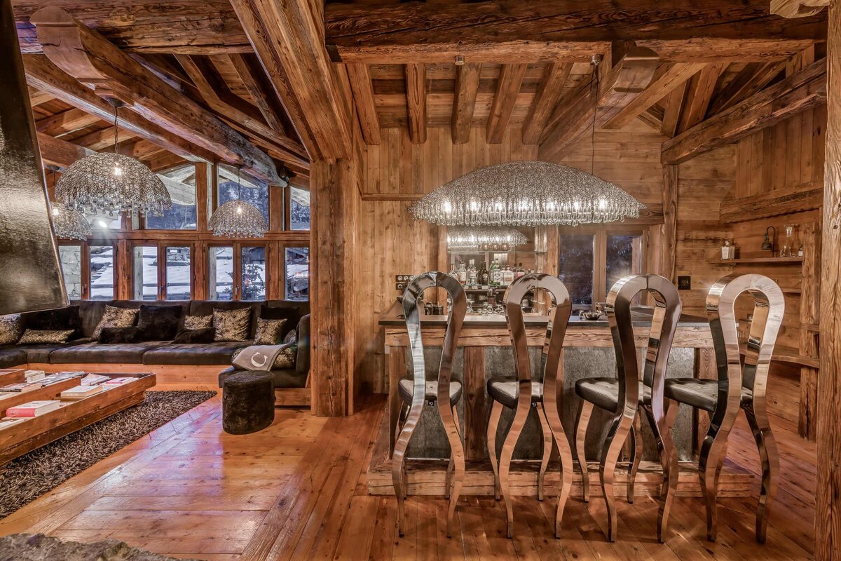 Rustic yet luxurious wooden chalet interior featuring a home bar with unique wavy chrome stools, a grand chandelier, and a cozy lounge.