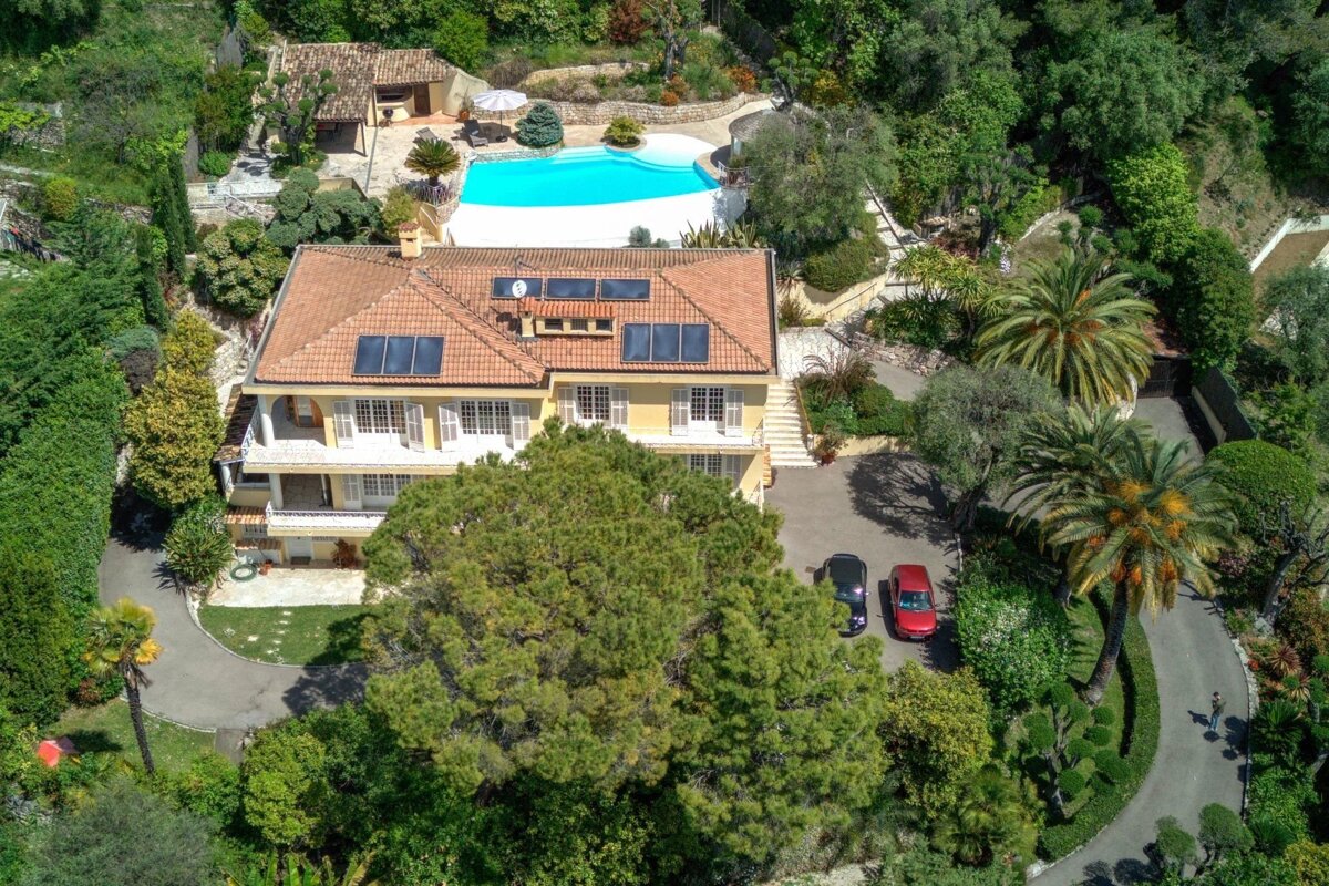 An aerial view of a large yellow villa with a tiled roof, featuring a backyard pool and surrounded by lush green trees.