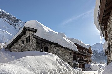 Snow-covered stone alpine chalets in a mountainous winter landscape under a clear blue sky.