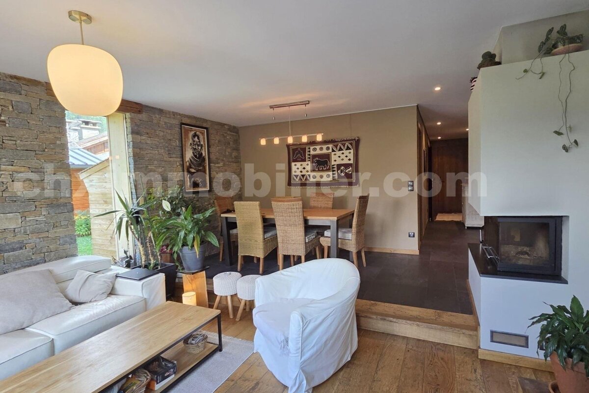 Rustic-modern open concept living/dining room with stone accents, white sofa, wooden table, fireplace, and plants.