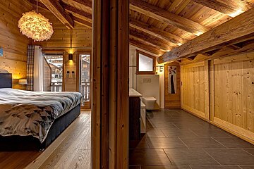 Inviting wood-paneled chalet bedroom with an en-suite bathroom, featuring exposed beams, warm lighting, and a balcony view of a snowy town.