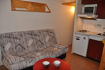 A cozy studio apartment with a patterned sofa, a red table with cups, and a compact kitchenette including a microwave, stove, and fridge.