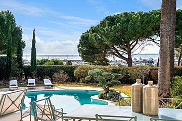 A beautiful outdoor scene with a swimming pool in a lush garden, featuring trees, hedges, and a distant marina filled with boats by the sea.