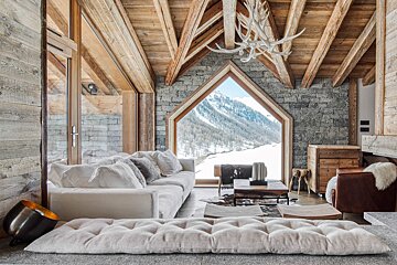 Rustic chalet living room with wooden beams, stone walls, and a large triangular window revealing a breathtaking snow-covered mountain landscape.