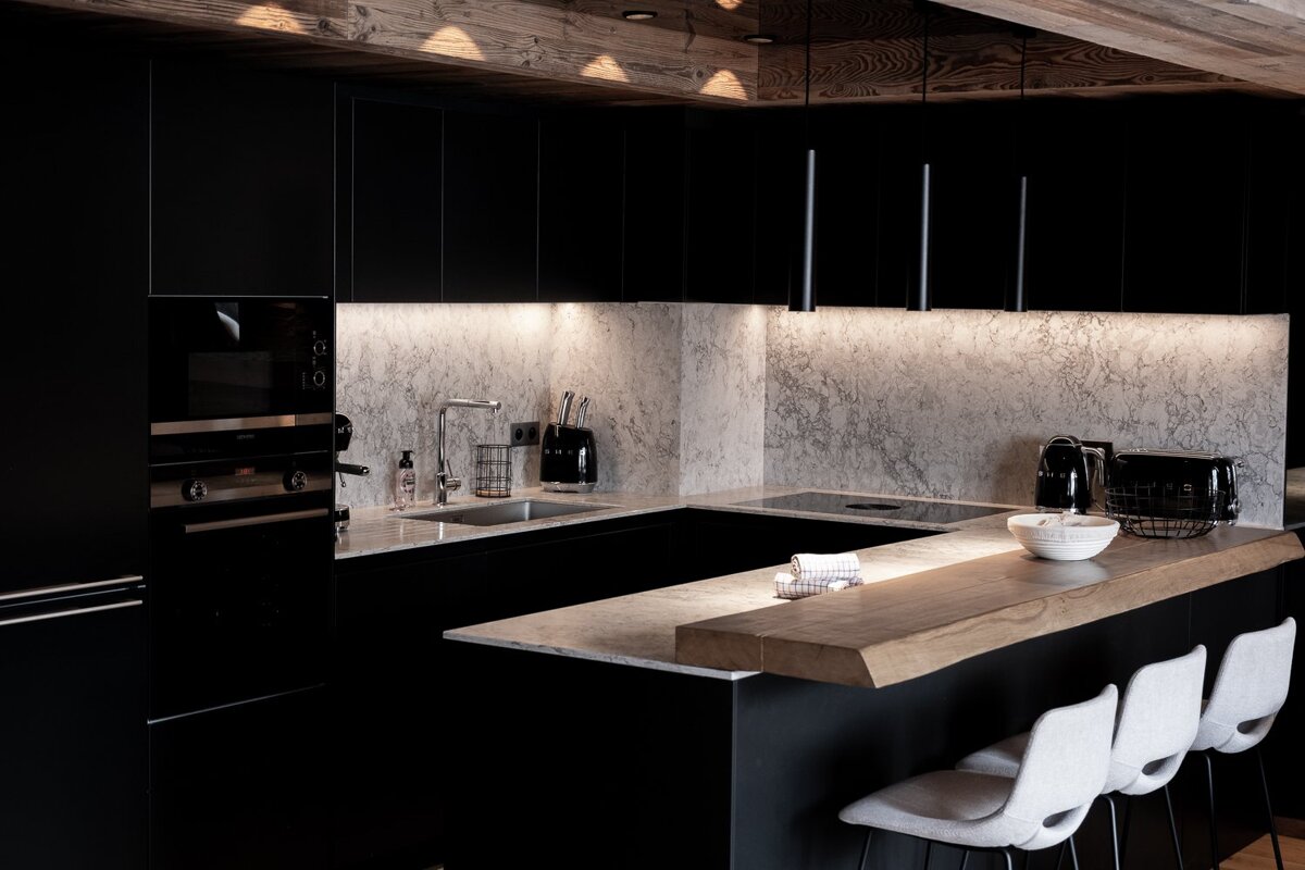 A sleek, modern kitchen featuring matte black cabinetry, marble countertops & backsplash, and a warm wooden breakfast bar with stools.