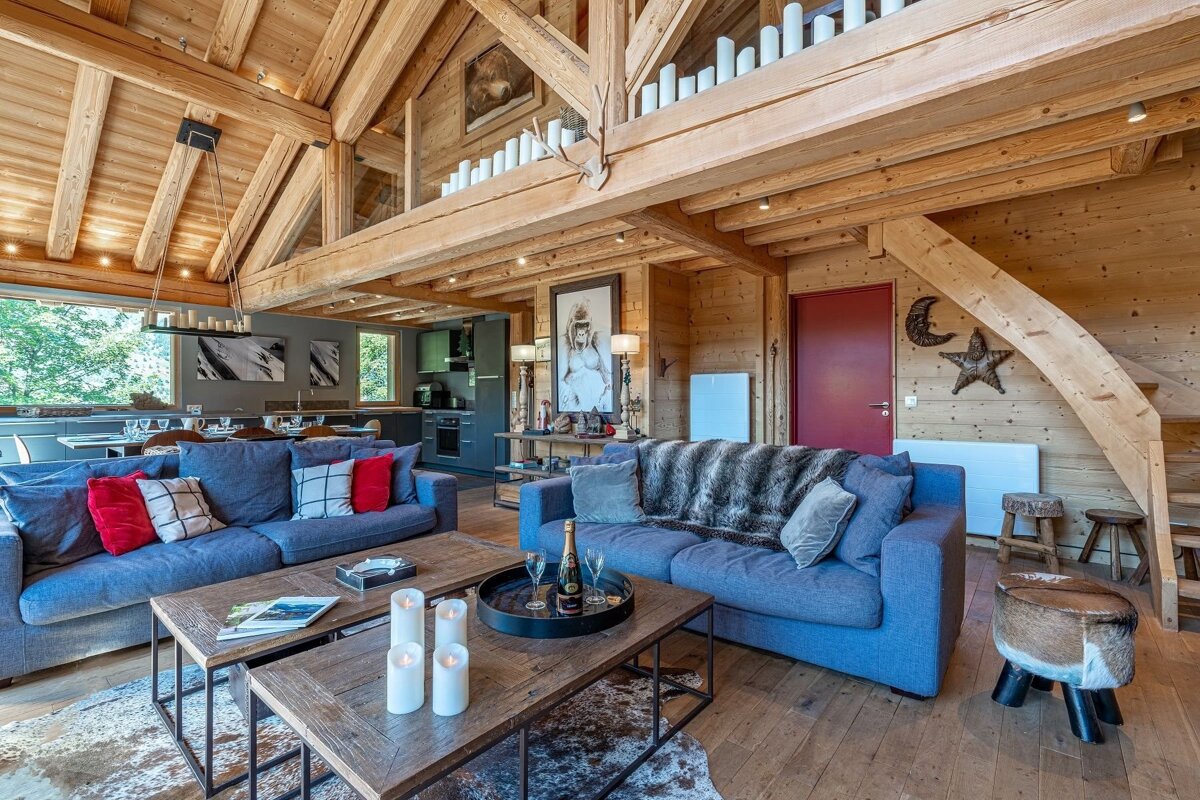 A spacious, rustic-chic chalet interior with exposed wooden beams, blue sofas, and modern kitchen. Cozy mountain retreat vibes.