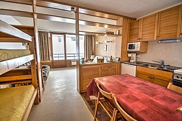 A compact, wood-paneled apartment interior featuring bunk beds, a kitchen with a dining table, and a living space leading to a balcony.