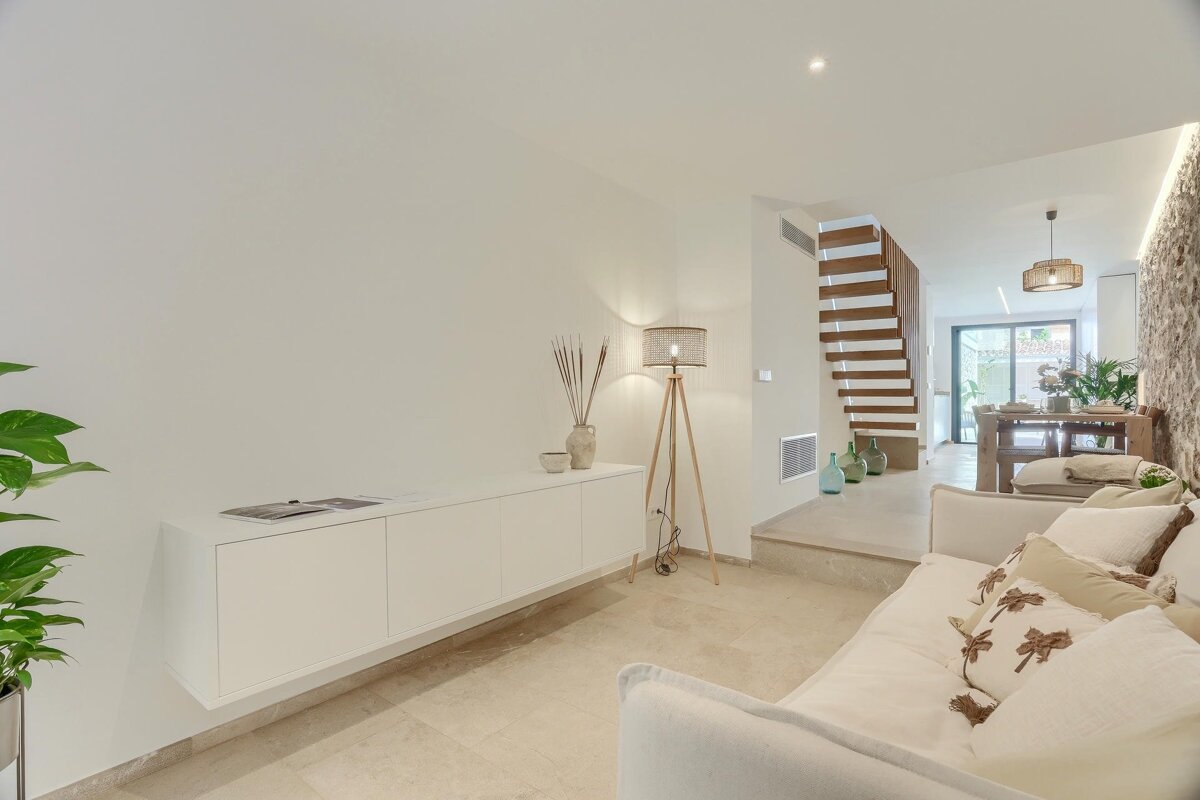 A bright, open-plan living room with a white sofa, floating cabinet, and wooden staircase. Light stone floors lead to a dining area with a textured stone wall.