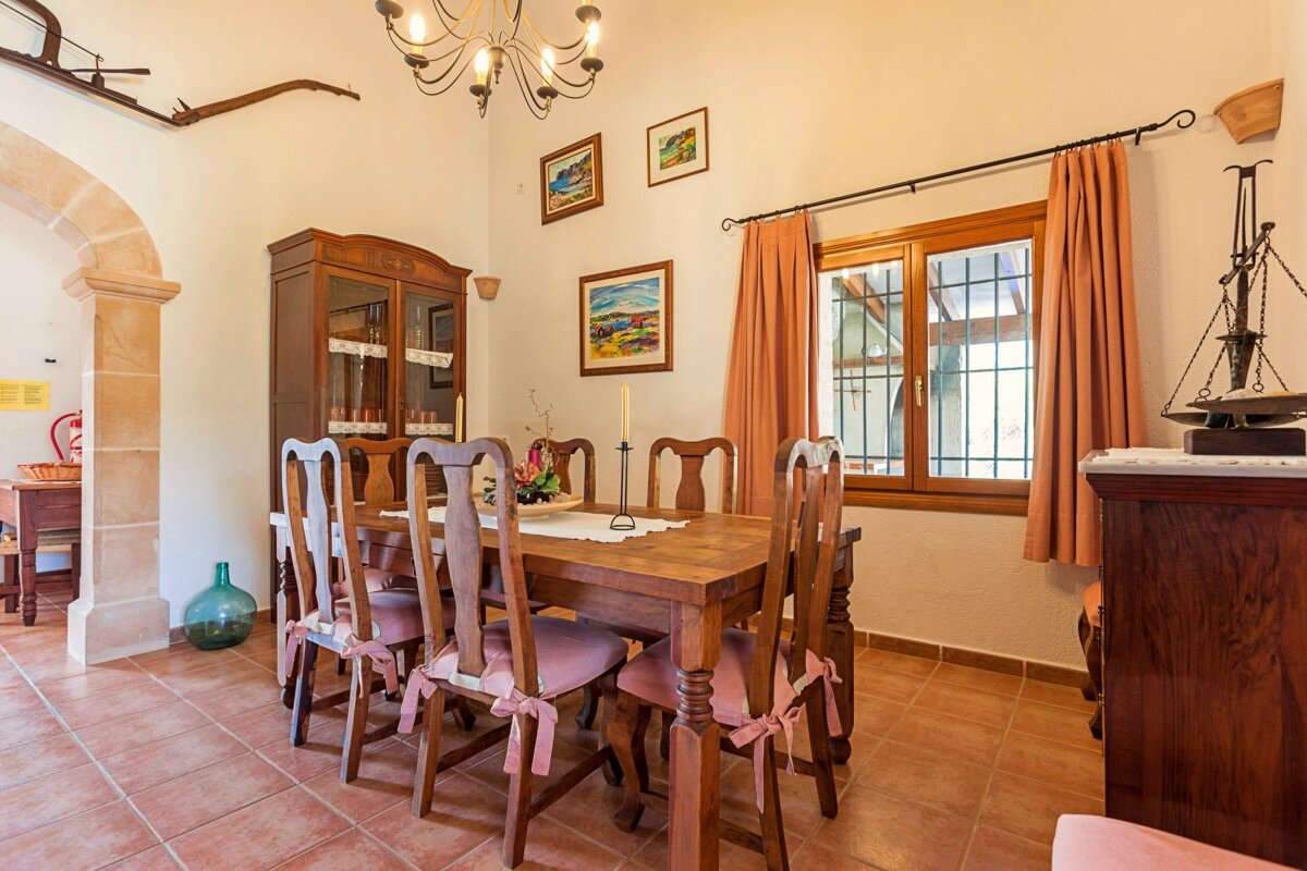 A rustic dining room with a large wooden table, chairs with pink cushions, a cabinet, and a window with peach curtains. Tiled floor and archway.