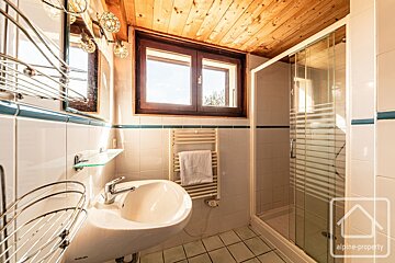 A bright, cozy bathroom featuring white tiles with blue trim, a warm wooden ceiling, a sink, mirror, window, towel warmer, and a glass shower cabin.