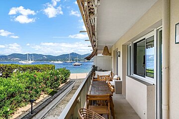 An apartment balcony with outdoor dining, overlooking a beautiful beach, blue sea with boats, and distant mountains.
