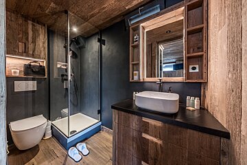 Chic rustic bathroom featuring a glass shower, toilet, vessel sink, and illuminated mirror against dark walls and rich wood textures.