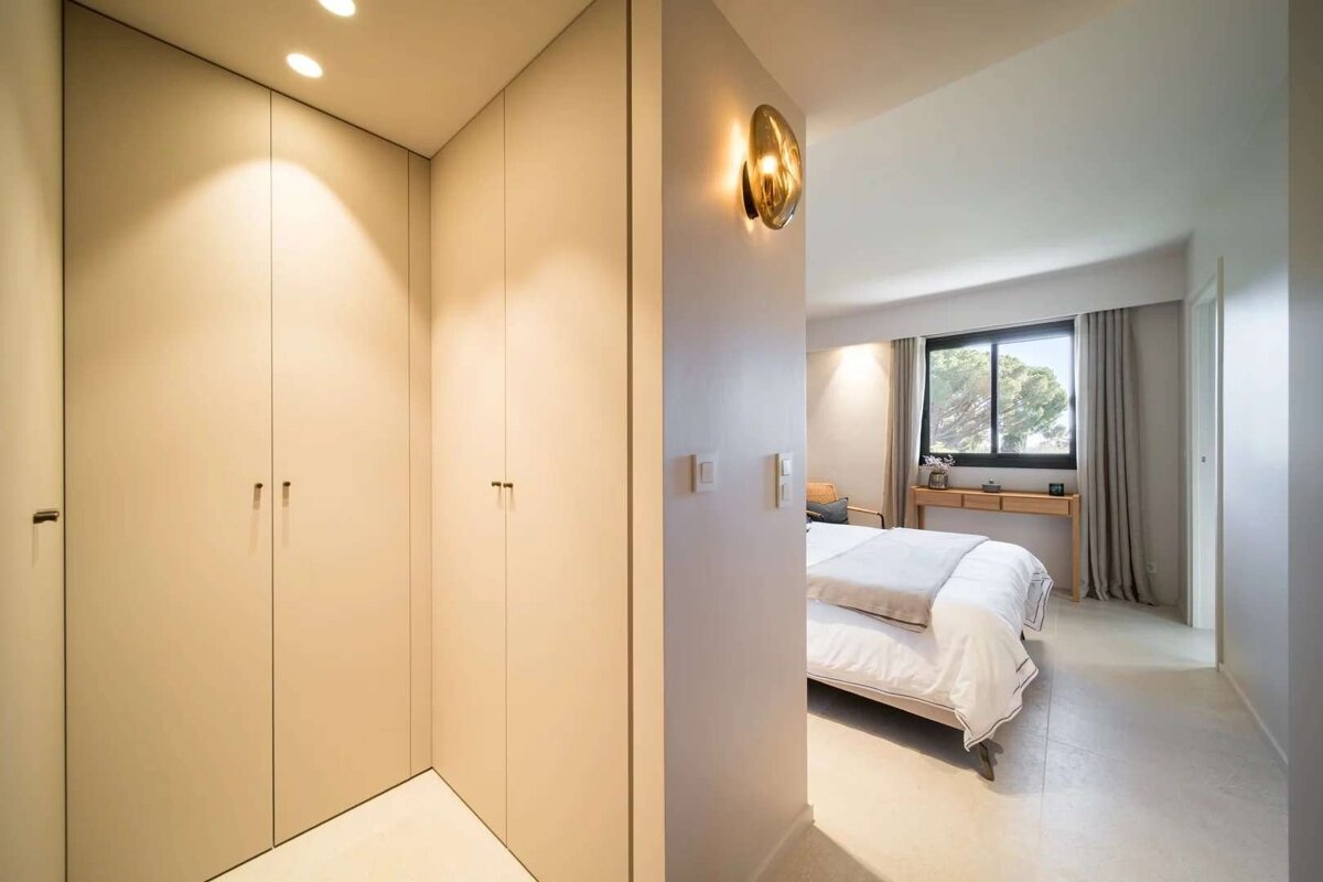 A bright, modern hallway with built-in light wardrobes and warm lighting, leading to a bedroom with a bed and large window.