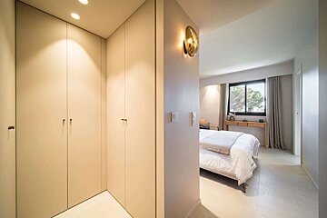 A bright, modern hallway with built-in light wardrobes and warm lighting, leading to a bedroom with a bed and large window.