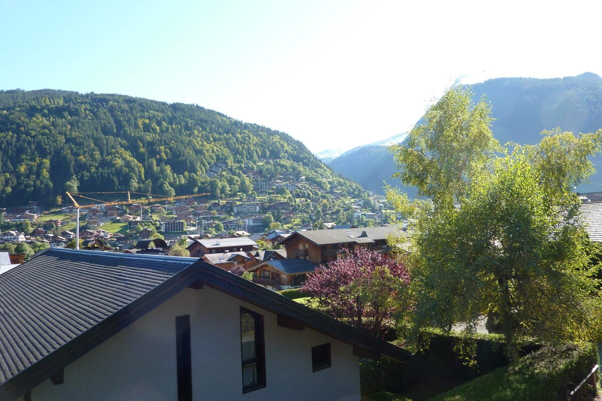 A panoramic view of a mountain village nestled in lush green hills under a clear sky. Traditional houses dot the landscape, with a crane visible on the left.