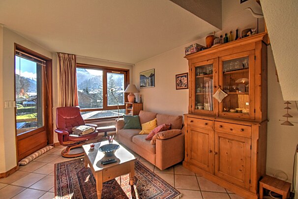 A cozy, sunny living room with a mountain view through large windows. Features a pink sofa, red armchair, and a rustic wooden cabinet on tiled floors.