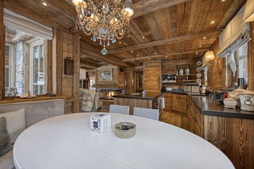 Cozy, rustic wooden chalet interior with a white oval dining table, crystal chandelier, kitchen, and a warm fireplace in the background.
