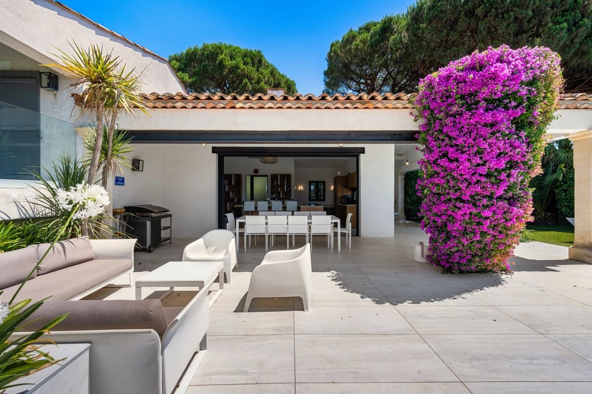 A chic villa patio with modern outdoor furniture and a striking column wrapped in vibrant purple bougainvillea under a clear blue sky.
