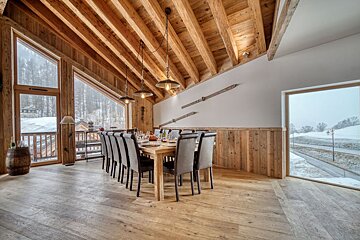 Rustic modern dining room with wood ceiling, walls, and floor. Large windows overlook a snowy landscape. Long table set for dinner with vintage skis on the wall.