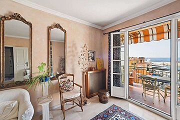 A cozy room with peach walls, ornate mirrors, and vintage furniture. French doors open to a balcony with an orange awning, revealing a scenic view of a seaside town and port.