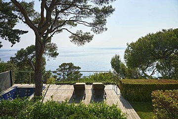 A wooden deck with two loungers and a jacuzzi overlooks a calm blue sea, framed by lush trees and greenery on a sunny day.