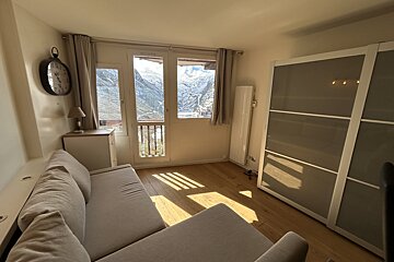 A sunlit room with wooden floors, a gray sofa, a large wardrobe, and glass doors opening to snowy mountain views.