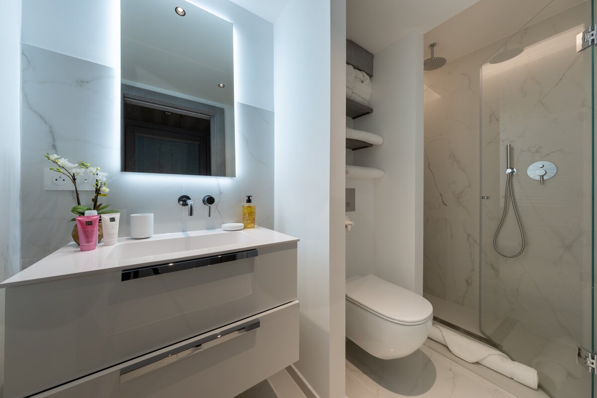 Sleek modern bathroom featuring a white vanity, illuminated mirror, wall-mounted toilet, and a glass-enclosed rain shower.
