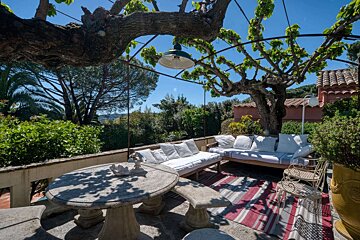 Sunny outdoor patio with a stone table, white cushioned sofas, and a large tree canopy providing shade over lush greenery and blue sky.