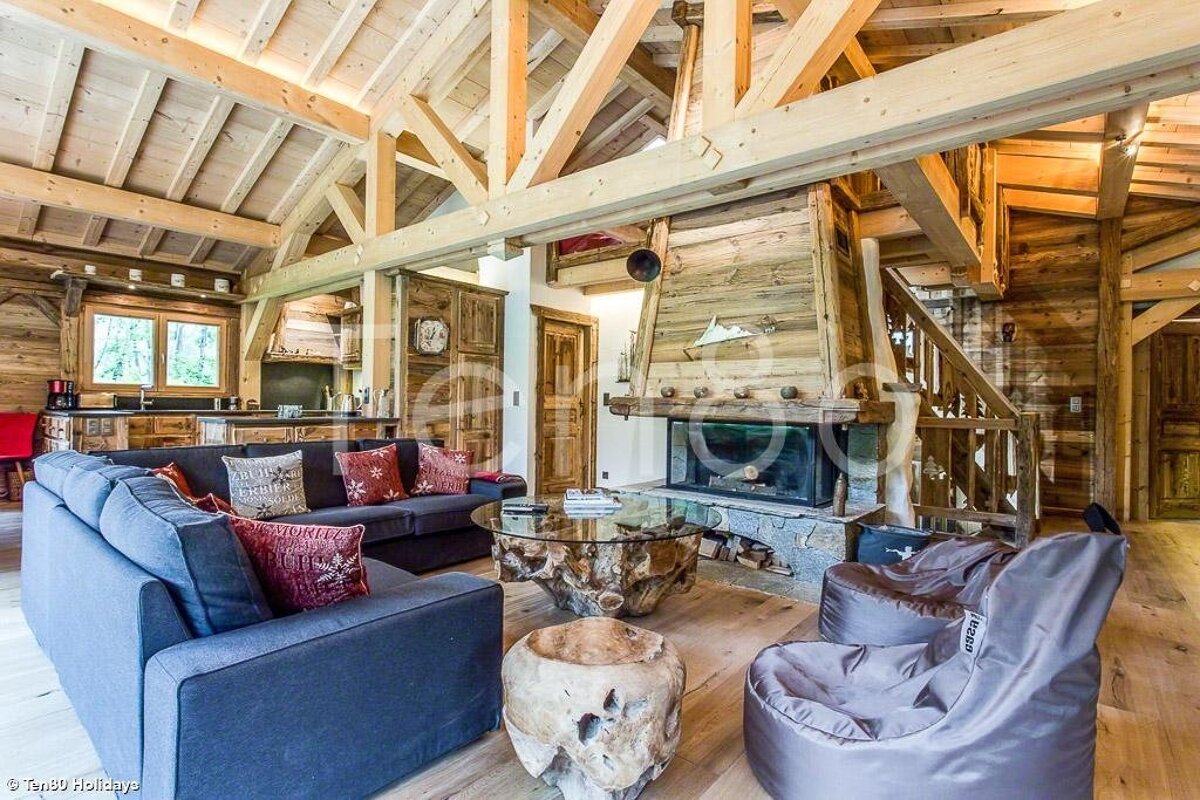 Cozy alpine chalet living room with exposed wood beams, a stone fireplace, grey sofas, and rustic wood-stump tables.