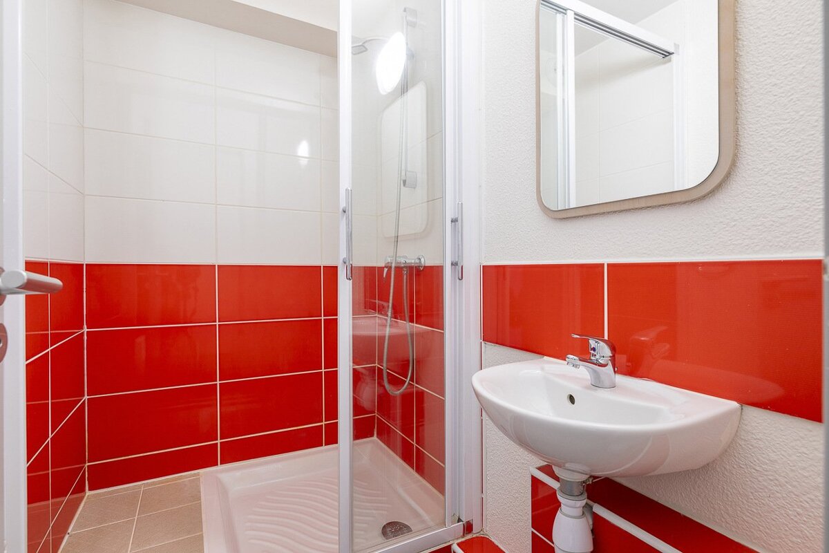 A bright bathroom with white and red tiled walls, featuring a glass-enclosed shower, a wall-mounted sink, and a mirror.