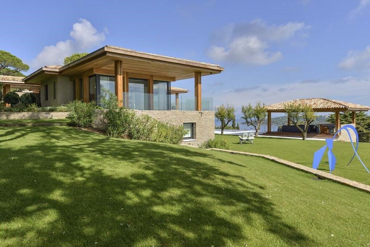 Modern villa with a large green lawn, blue abstract sculpture, and seaside gazebo under a bright sky.