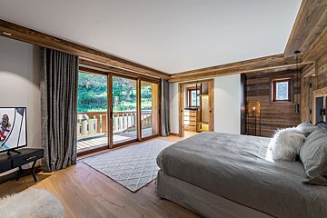 Cozy chalet bedroom with wooden walls, a large grey bed, and sliding glass doors opening onto a scenic balcony.