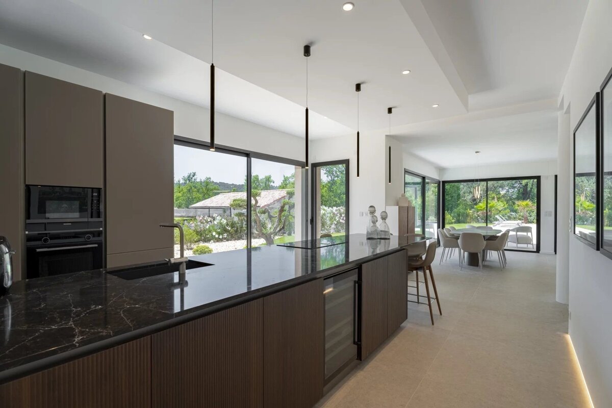 Sleek, modern kitchen with a dark marble island, integrated appliances, and an open layout leading to a bright dining area and lush outdoor view.