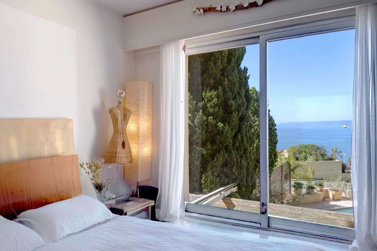 A serene bedroom with a white bed and large sliding doors opening to a beautiful ocean view, tall green trees, and distant coast.