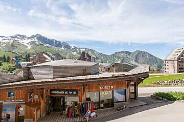 Mountain resort village with wooden shops (Quiksilver, Roxy, Skiset) and buildings. Snow-capped mountains and green hills under a bright sky.