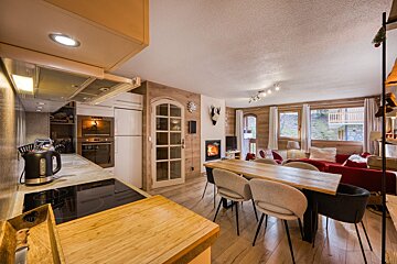 Warm, open-plan chalet living space featuring a modern kitchen, dining area, and living room with a fireplace, wooden walls, and comfy red sofa.