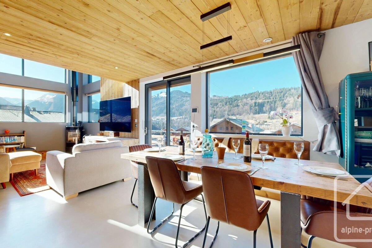 Sunny, modern ski chalet interior with wood ceilings, a set dining table, and large windows revealing panoramic mountain views.