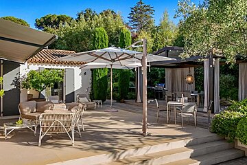 Spacious outdoor patio with lounge and covered dining areas, a large umbrella, and lush greenery under a clear sky.