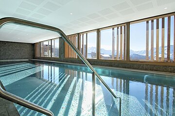 A modern indoor swimming pool features large windows offering panoramic views of snowy mountains, with sunlight dappling on the water's surface.