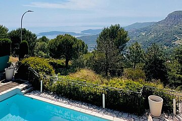 A blue swimming pool with a scenic view of lush green hills, distant mountains, and the sea under a clear sky.