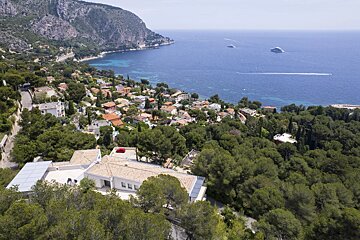 Aerial view of a lush coastal hillside with houses nestled in trees, overlooking a blue sea with distant yachts.