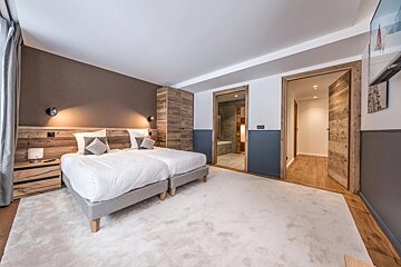 Modern bedroom with twin beds, wooden accents, white rug, and an en-suite bathroom. Warm lighting and two-tone walls create a cozy atmosphere.