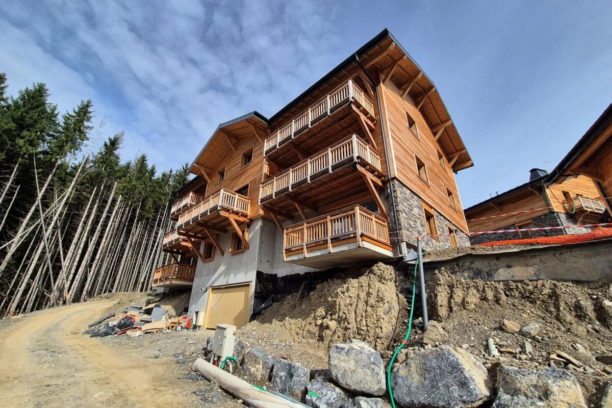 A multi-story wooden chalet built on a rocky hillside next to a pine forest under a blue sky.