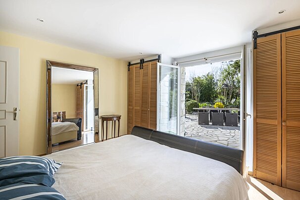 Bright bedroom with a large bed, wooden shutter closets, and open glass doors leading to a sunny stone patio.