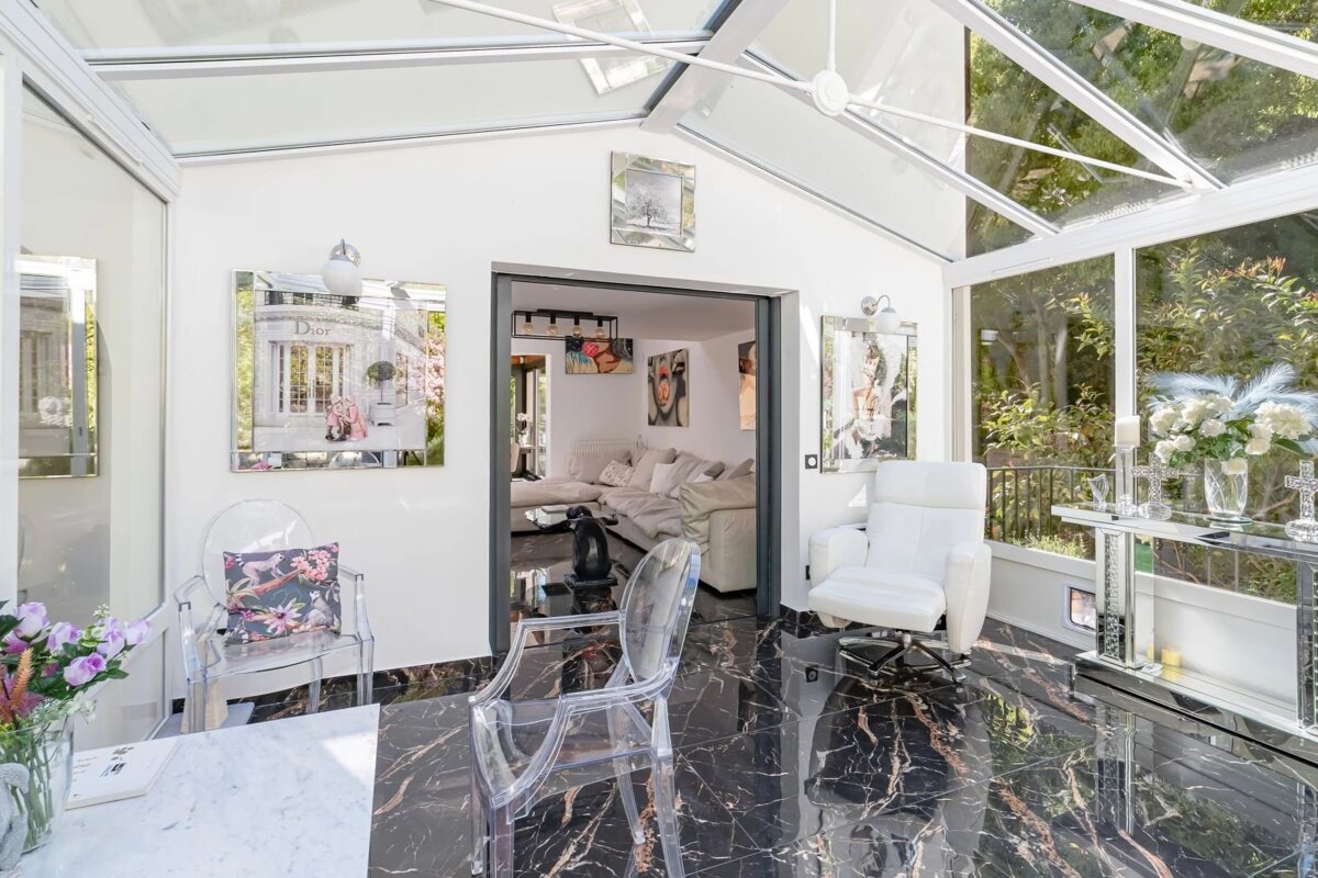 Bright sunroom with black marble floors, a glass roof, clear acrylic chairs, and a white armchair opening into a living room.