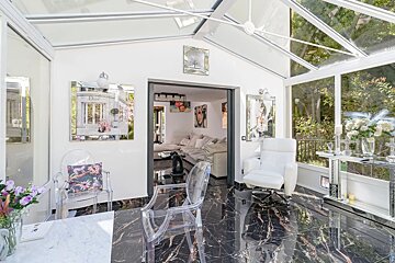 Bright sunroom with black marble floors, a glass roof, clear acrylic chairs, and a white armchair opening into a living room.