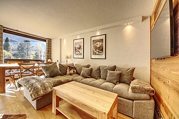 A cozy living room features a large L-shaped sofa, wooden furniture, and a dining area by a panoramic window with mountain views. Ski posters adorn the wall.