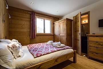 Cozy rustic bedroom with wood-paneled walls, a bed with a patterned red blanket, wooden furniture, and an open door to a bathroom.