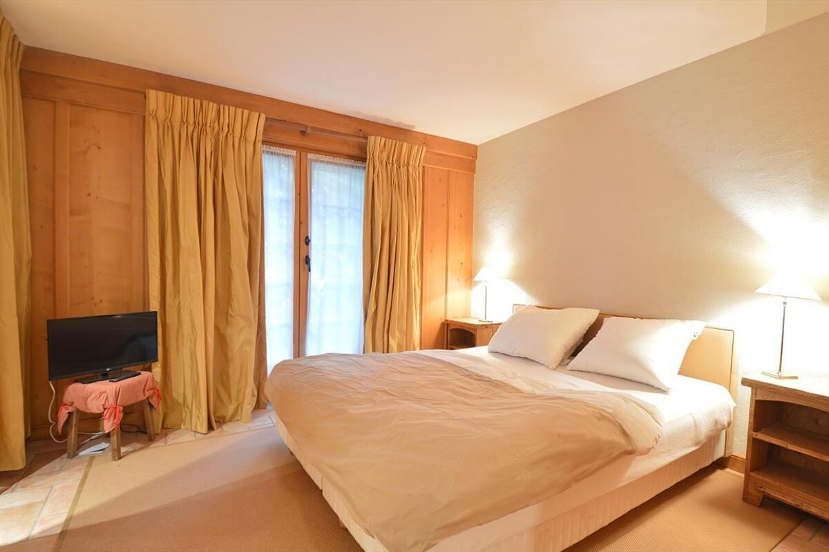 A cozy bedroom with a double bed, light bedding, wood-paneled walls, and yellow curtains. A TV is on the left, and lamps flank the bed.