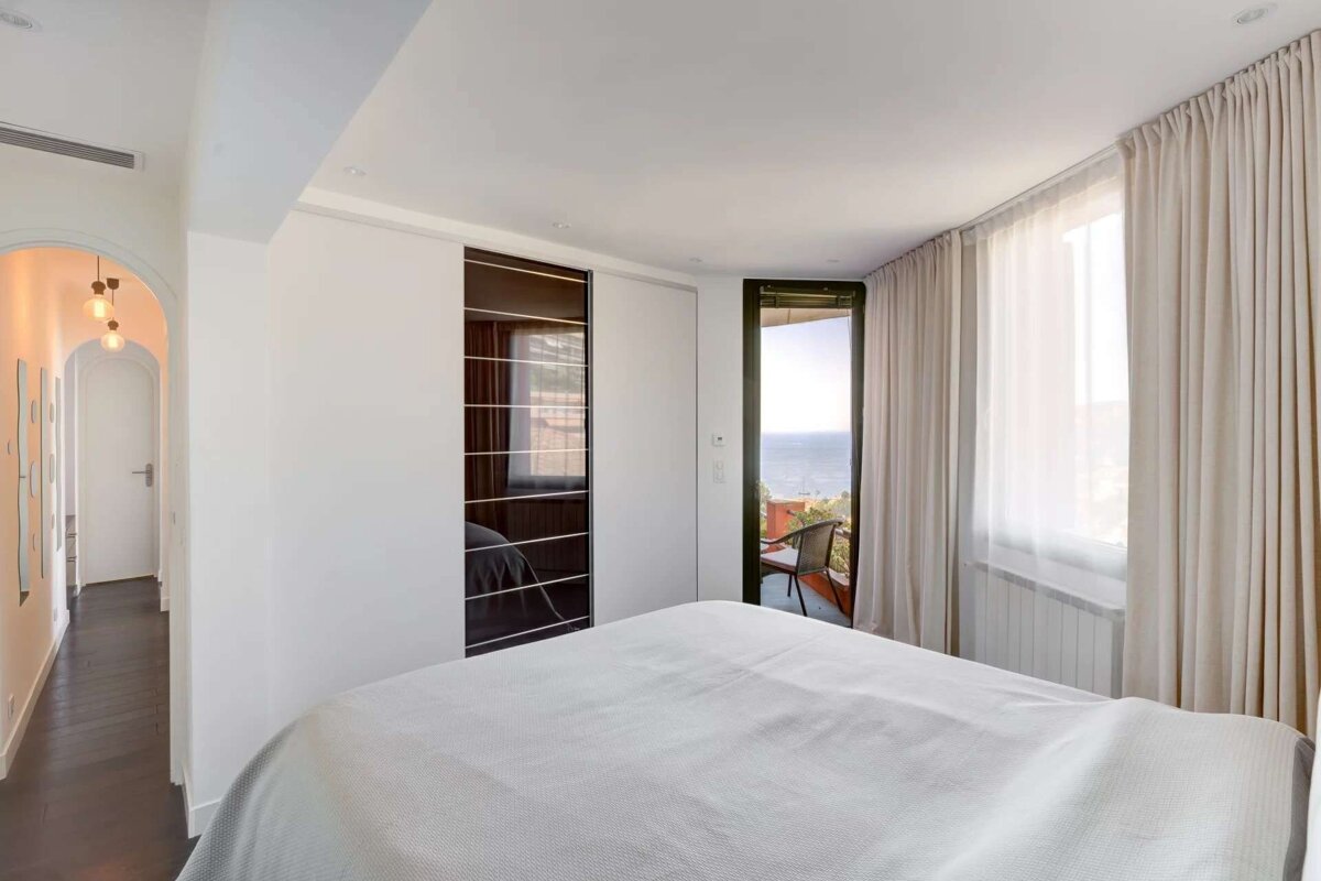 A modern bedroom with white bedding and a dark sliding closet opens to a balcony with an ocean view. An arched hallway is visible.